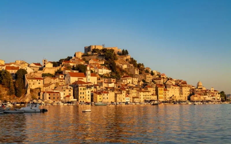 A picturesque view of Šibenik's historic waterfront at sunset, reflecting golden hues.