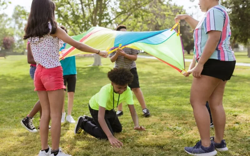 Group of children playing with a colorful parachute outdoors on a sunny day.