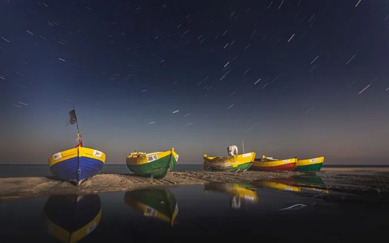 Colorful fishing boats on a beach under a starry sky in Jastarnia, Poland, at nighttime.