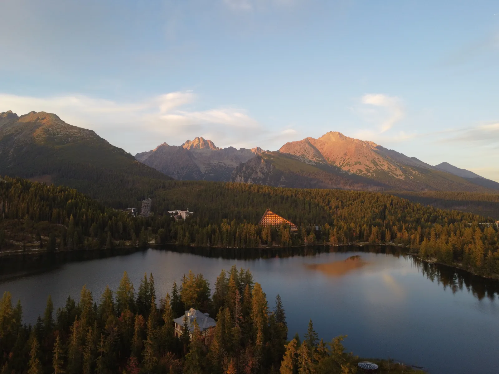 Evening alpenglow illuminates Vysoká and Končistá peaks in the High Tatras, viewed from a drone over Štrbské Pleso lake. Hotel Patria is visible on the far shore.