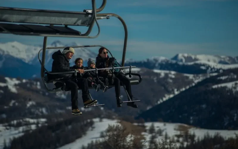 Family having fun on a ski lift with snow-covered mountains in the background during a sunny winter day.