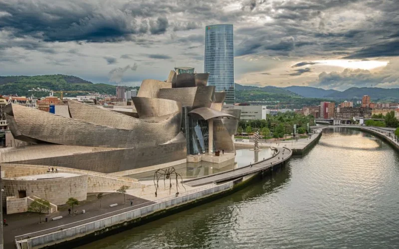 A striking view of the Guggenheim Museum Bilbao by the river under dramatic clouds, showcasing its iconic architecture.
