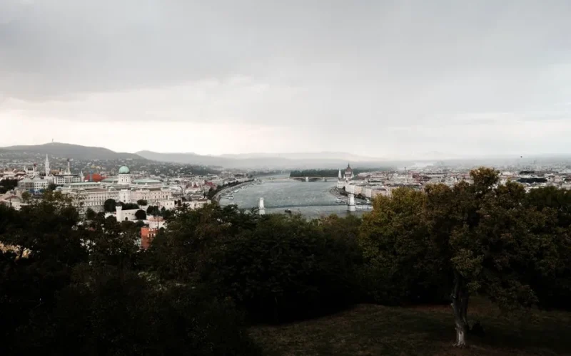 Scenic view of Budapest's cityscape with the Danube River and iconic landmarks under a cloudy sky.