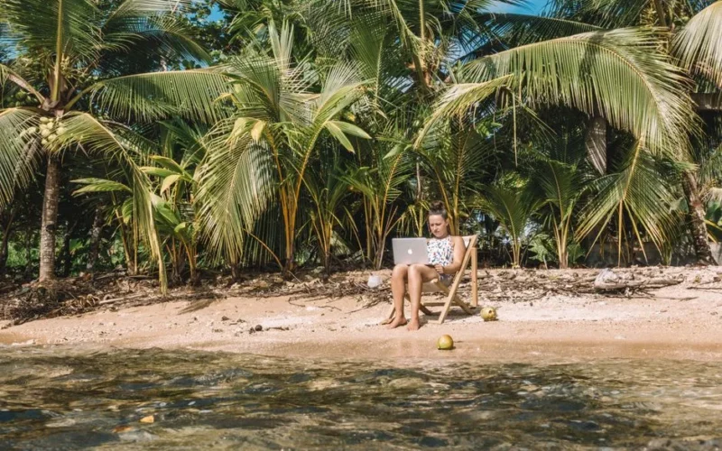 Woman working remotely with a laptop on a tropical beach in Panama surrounded by lush green palm trees.