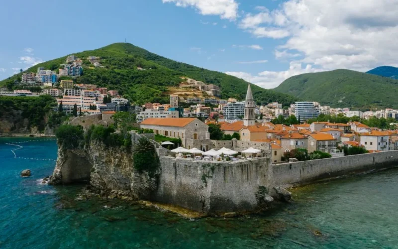 Aerial view of a historic town on the Adriatic coast, featuring old stone walls and lush green mountains.
