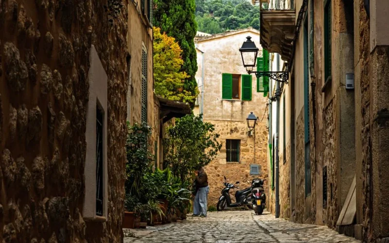 Quaint cobblestone street with rustic walls, greenery, and parked scooters in a picturesque European village.