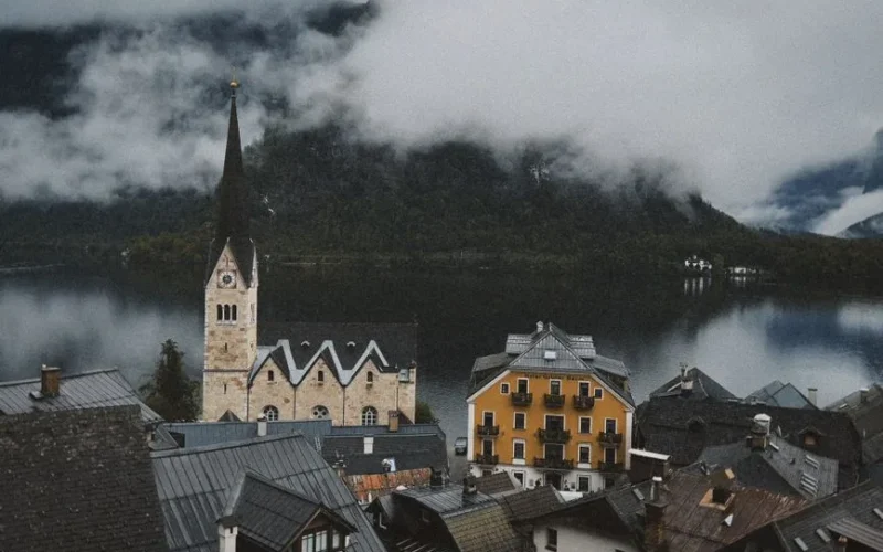 Scenic view of Hallstatt village with iconic church and serene lake surrounded by misty mountains.