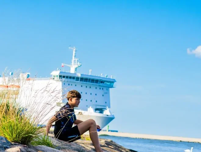 Young boy sitting on rocks by the sea with a ferry in the background under a clear blue sky.