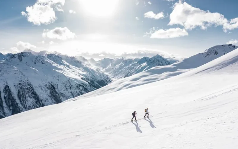 Two skiers climbing a sunlit snowy mountain slope in Ischgl, Austria, during winter.