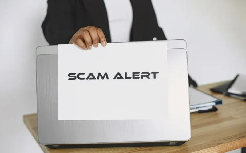 Business person holding a scam alert sign over a laptop, warning against online fraud.