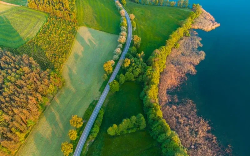 Vibrant aerial view of a winding road through lush green Kaszuby countryside beside a serene lake.