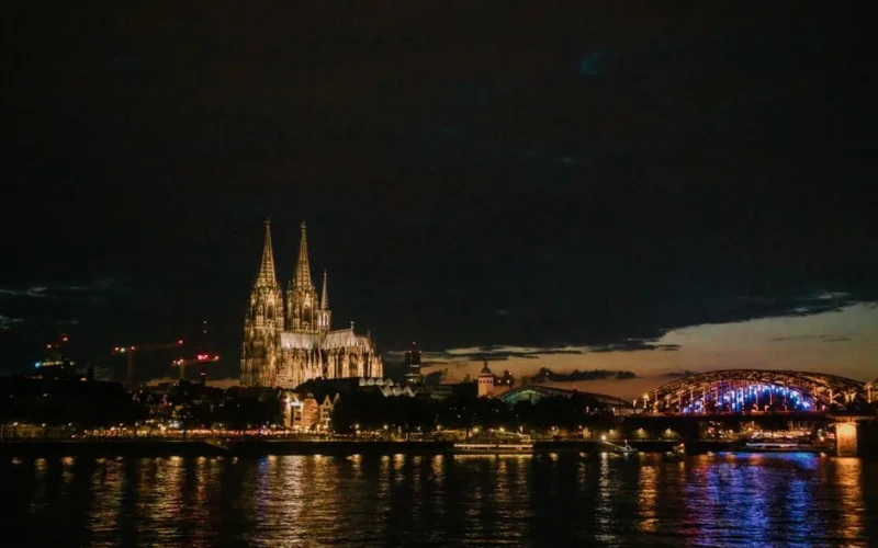 View of Cologne Cathedral and Hohenzollern Bridge illuminated at night, reflecting in the Rhine River