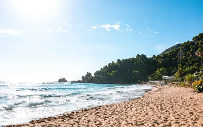 Discover a serene beach on Corfu, Greece, basking in the warm summer sun with lush greenery and clear blue waves.