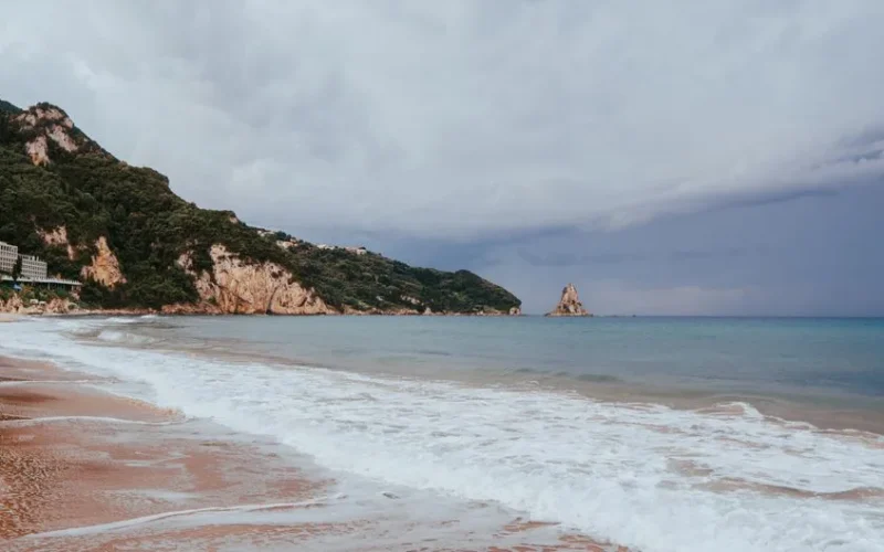 A serene seascape of Corfu's beach with golden sands, turquoise waters, and lush cliffs under a cloudy sky.