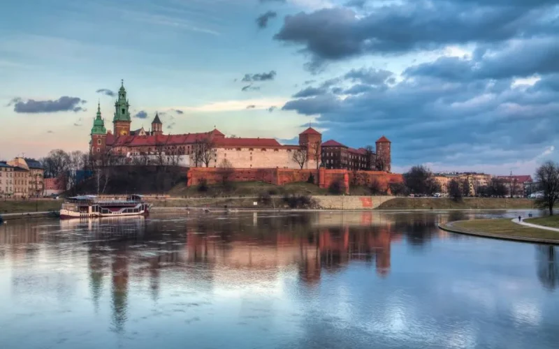 Serene view of Wawel Castle reflecting in the Vistula River at sunset in Kraków, Poland.