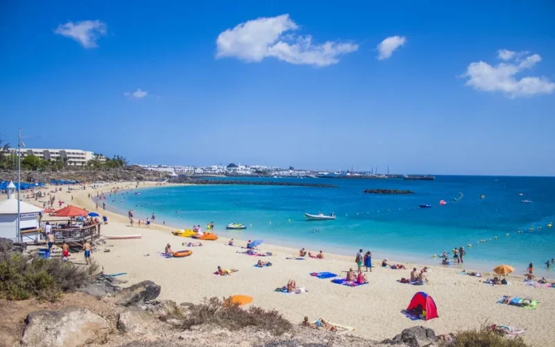 People enjoying a sunny day at Playa Blanca beach, Lanzarote with clear blue waters and golden sand.