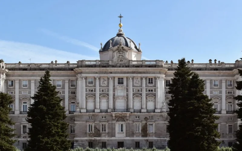 The grand facade of the Royal Palace of Madrid under a clear blue sky, framed by lush trees.