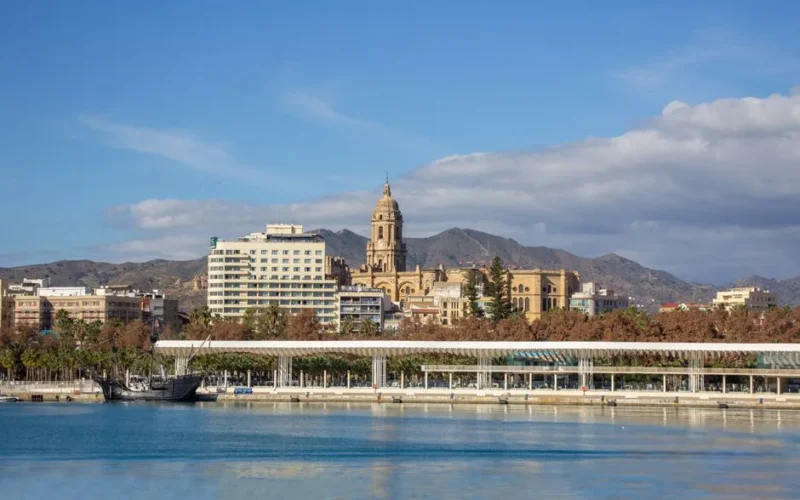Málaga skyline featuring the iconic cathedral, promenade, and calm waters under a clear sky.