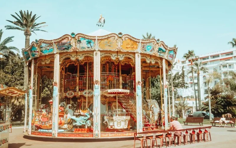 A colorful vintage carousel in a sunny amusement park with palm trees and vibrant decorations.