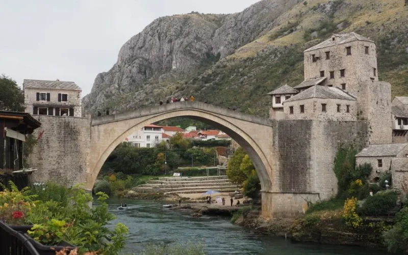 Scenic view of the historic Stari Most Bridge over the Neretva River in Mostar, surrounded by hills and buildings.