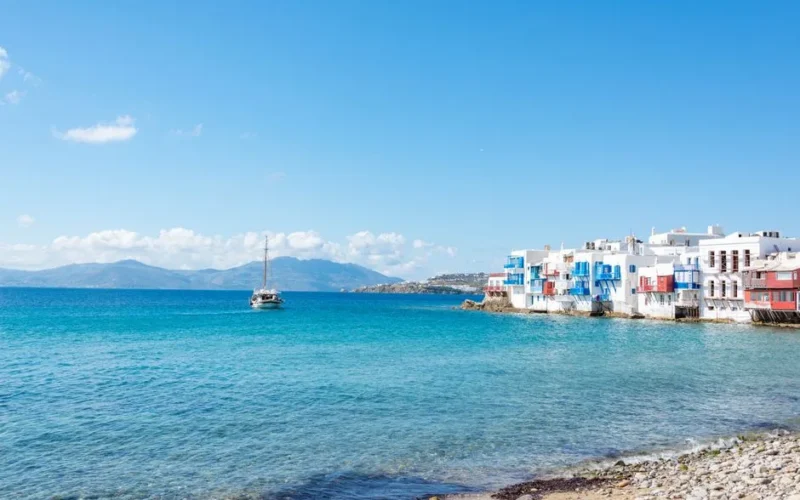 A stunning view of Mykonos with iconic white buildings and a sailboat on the turquoise water.