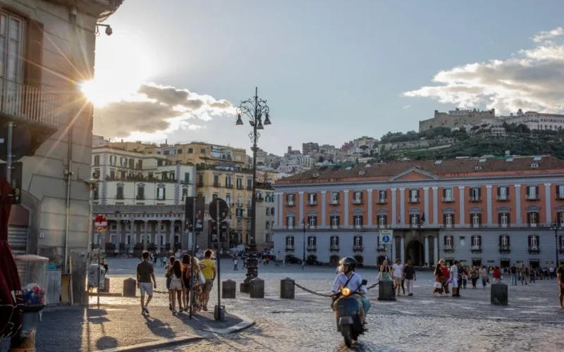 People walking and a scooter in a bustling piazza at sunset in Naples, Italy, showcasing historic buildings.