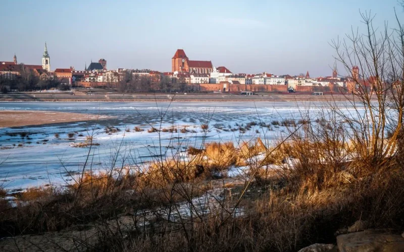 A scenic winter view of the historic Toruń cityscape across an ice-covered Vistula River.