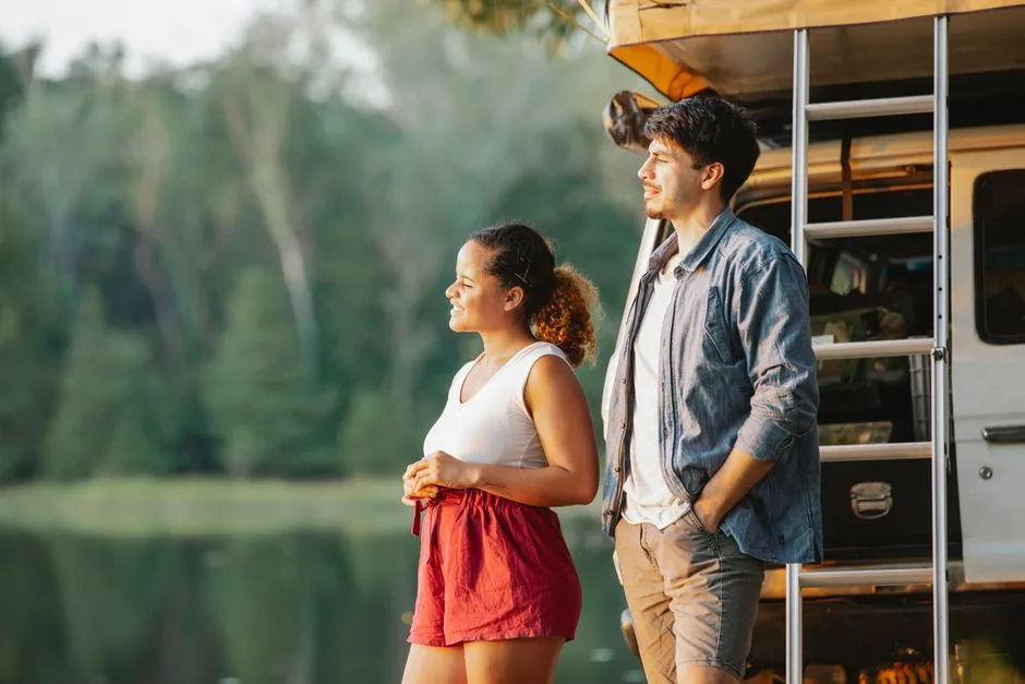 Side view of content young multiracial couple in casual outfits standing near SUV car on lake shore and admiring nature during road trip in nature at sunset