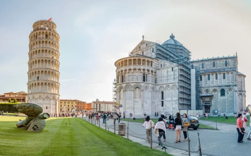Leaning Tower of Pisa and Pisa Cathedral, Italy, with visitors in Piazza del Duomo under a clear sky.