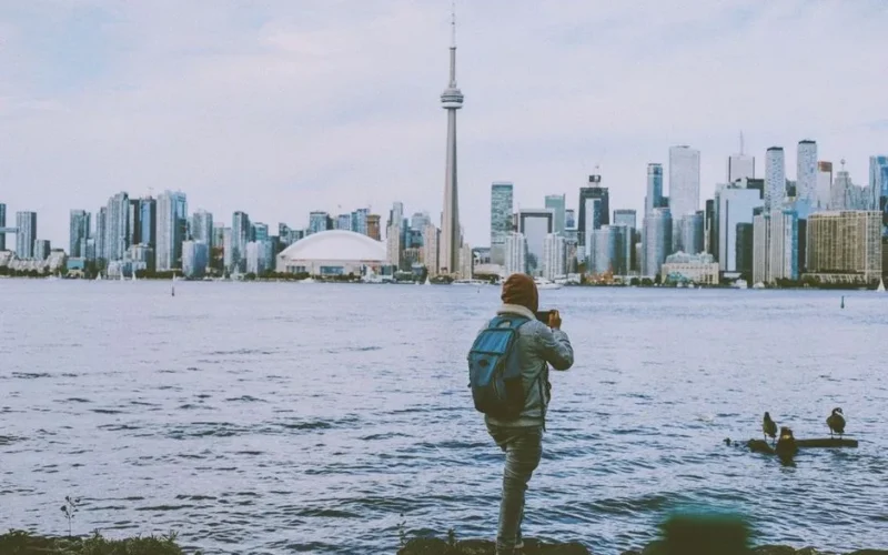 A backpacker taking photos of the Toronto skyline from across the water, featuring the CN Tower.