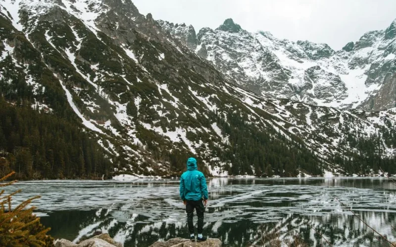 A solitary hiker gazes across a frozen mountain lake in winter, surrounded by snow-capped peaks.