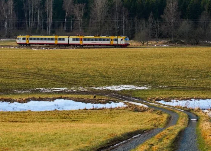 A scenic view of a passenger train traveling through a rural meadow with snow patches.