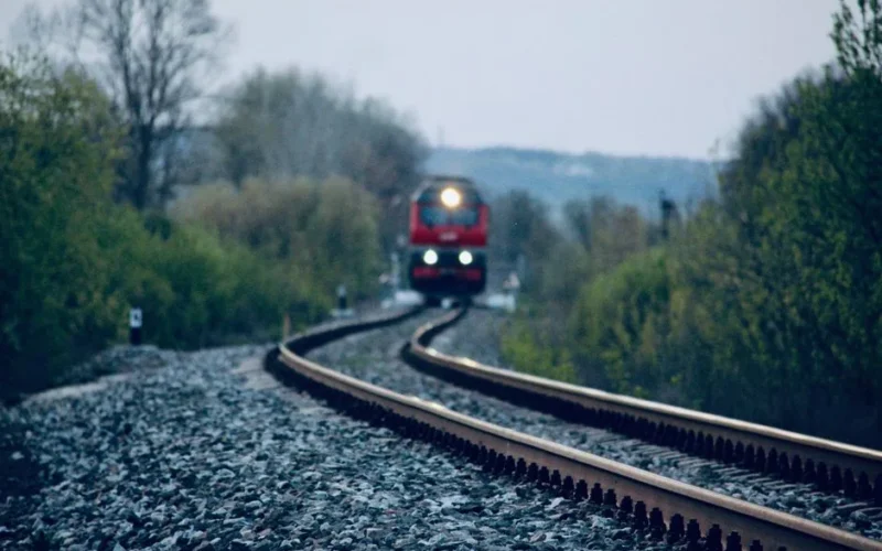 A red train approaching on a curved railway track with lush greenery during dusk.