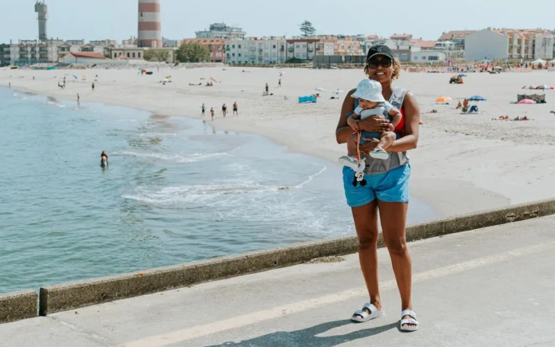 A mother holding her baby at a sunny beach near a lighthouse, capturing a joyful summer outing.