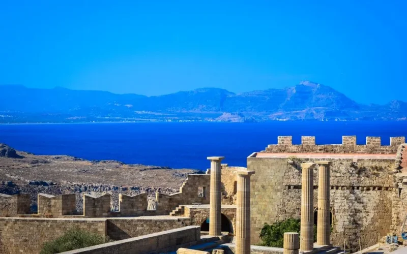 Scenic view of ancient Greek ruins with stone columns and walls against a vibrant blue Aegean Sea backdrop.