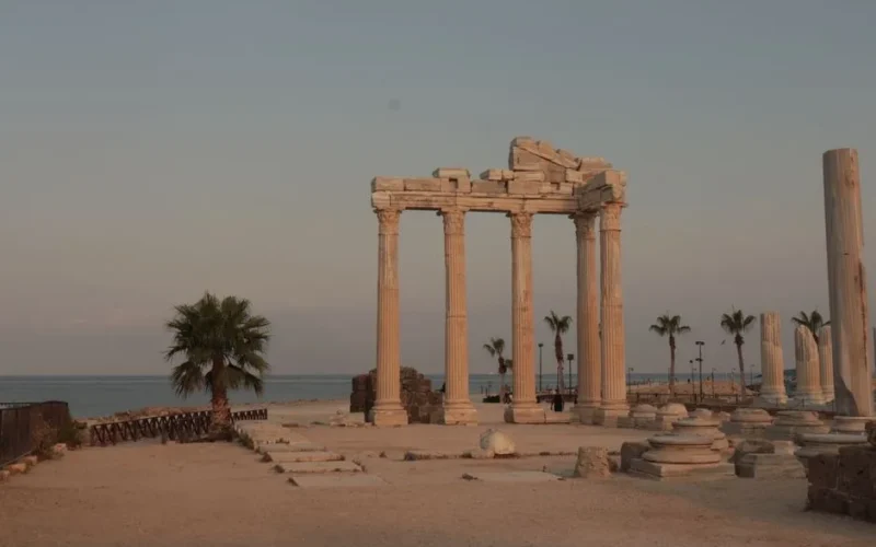 Scenic view of ancient Roman temple ruins by the sea at dusk, with palm trees.
