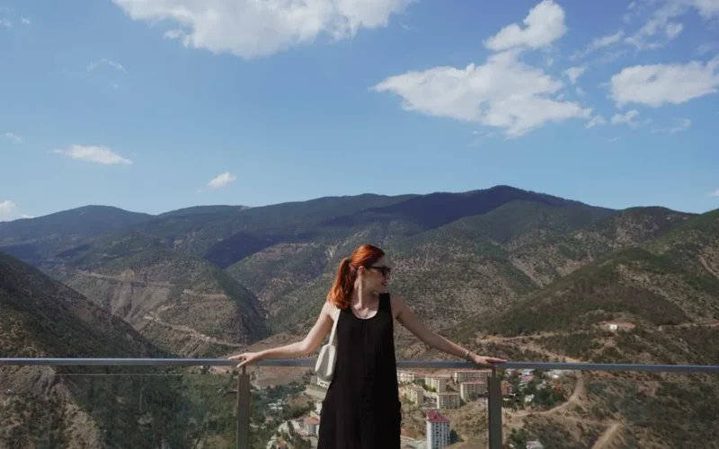 A woman admires the scenic mountain view from a glass terrace in Gümüşhane, Türkiye on a sunny day.