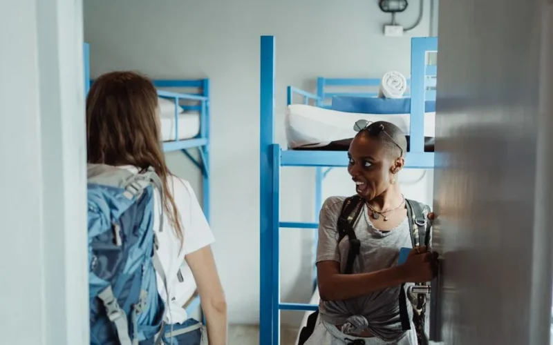 Two women with backpacks walking into a bright hostel dormitory with bunk beds, ready for adventure.