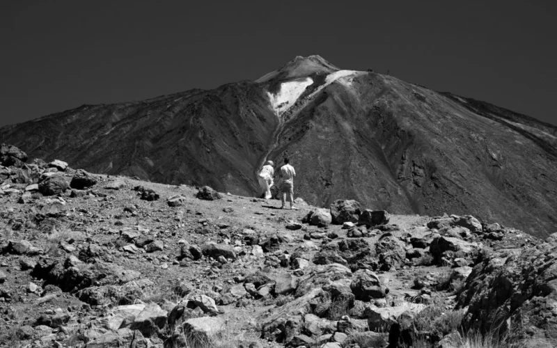 Two adults hike across volcanic terrain towards Pico del Teide, a popular attraction in Tenerife, Spain.