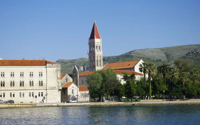 Scenic view of Trogir waterfront with the bell tower of St. Lawrence Cathedral against a mountainous backdrop.