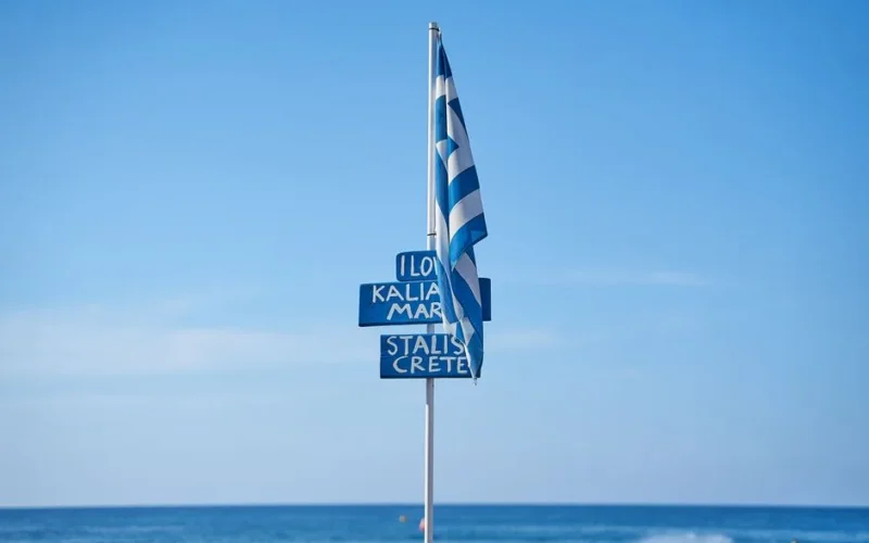 Greek flag flying at a beach in Crete, with blue sky and sea view.