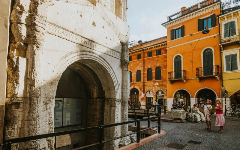 Explore a vibrant street in Verona, Italy, featuring a Roman arch and colorful historic architecture.
