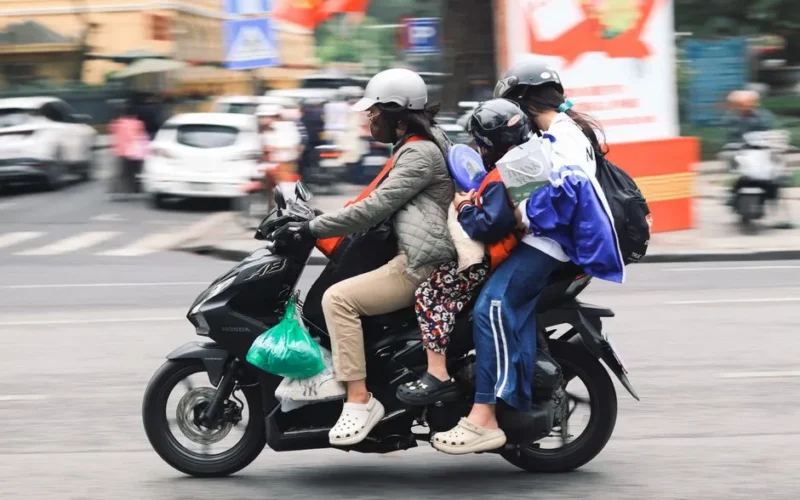 A family of four travels on a motorbike down a lively urban street, demonstrating local transportation methods.