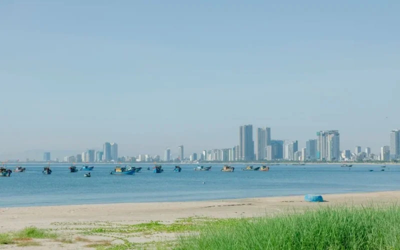 City skyline with boats on a calm ocean and sandy beach, under a clear blue sky.
