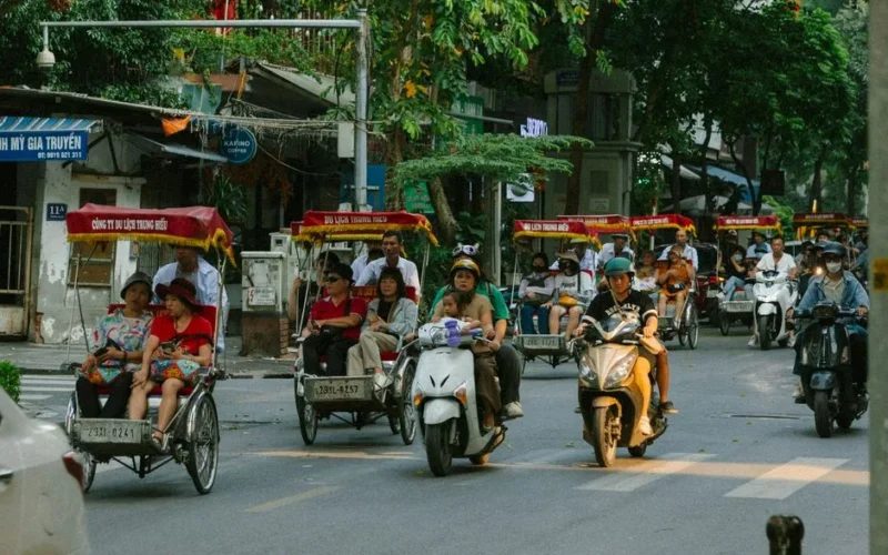 Vibrant city street featuring cyclos, motorbikes, and diverse people during the day.