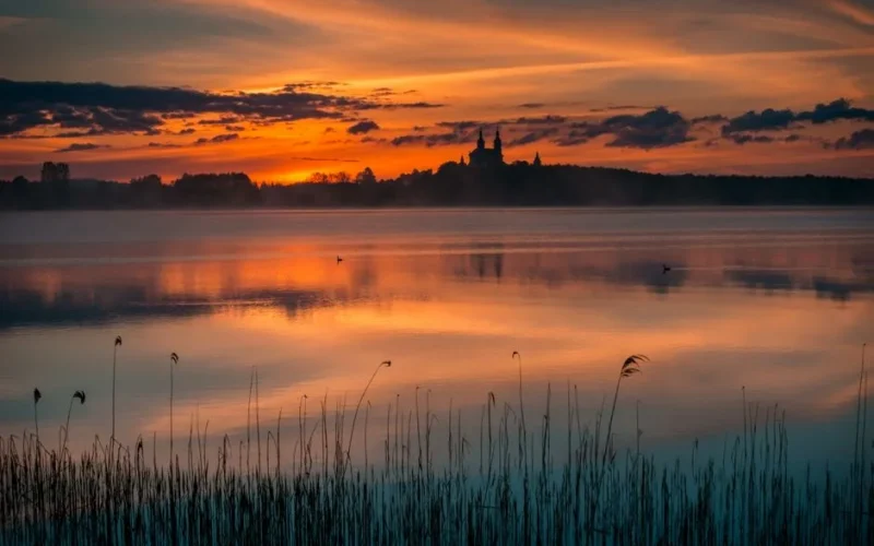 A tranquil scene of a sunset over a lake with reeds and a distant church silhouette, reflecting vibrant colors.