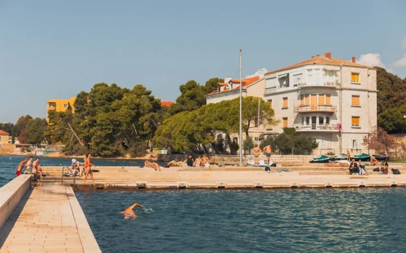 Vibrant seaside scene in Zadar, Croatia, showing people enjoying swimming and sunbathing under a sunny sky.