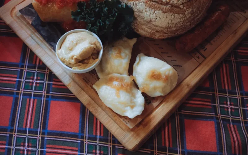 A wooden board with Polish pierogi, bread, and sausages on plaid tablecloth, showcasing authentic Polish cuisine.