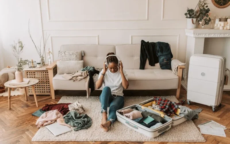 A woman packs a suitcase in her living room while listening to music, surrounded by clothes and a cozy atmosphere.
