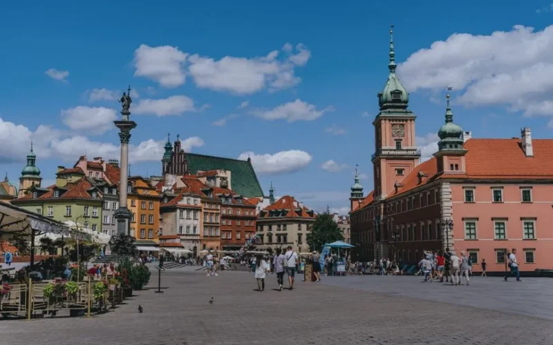 Vibrant scene of Warsaw's Old Town Square with tourists and iconic landmarks under a blue sky.